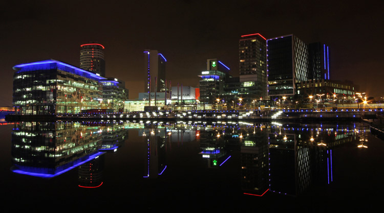 A night time photo of MediaCity, Salford, Manchester showing
            the neon lights reflecting against the water